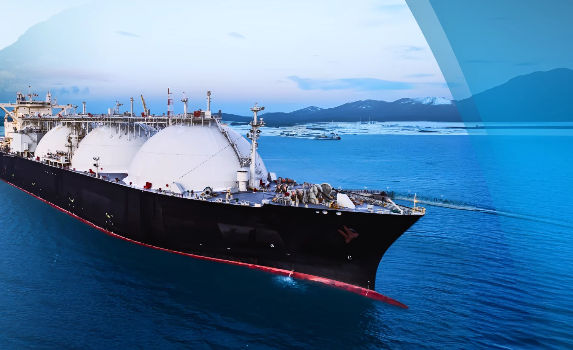 A large liquefied natural gas (LNG) tanker with spherical white storage tanks sails through calm blue water near a coastline with mountains in the background under a partly cloudy sky.
