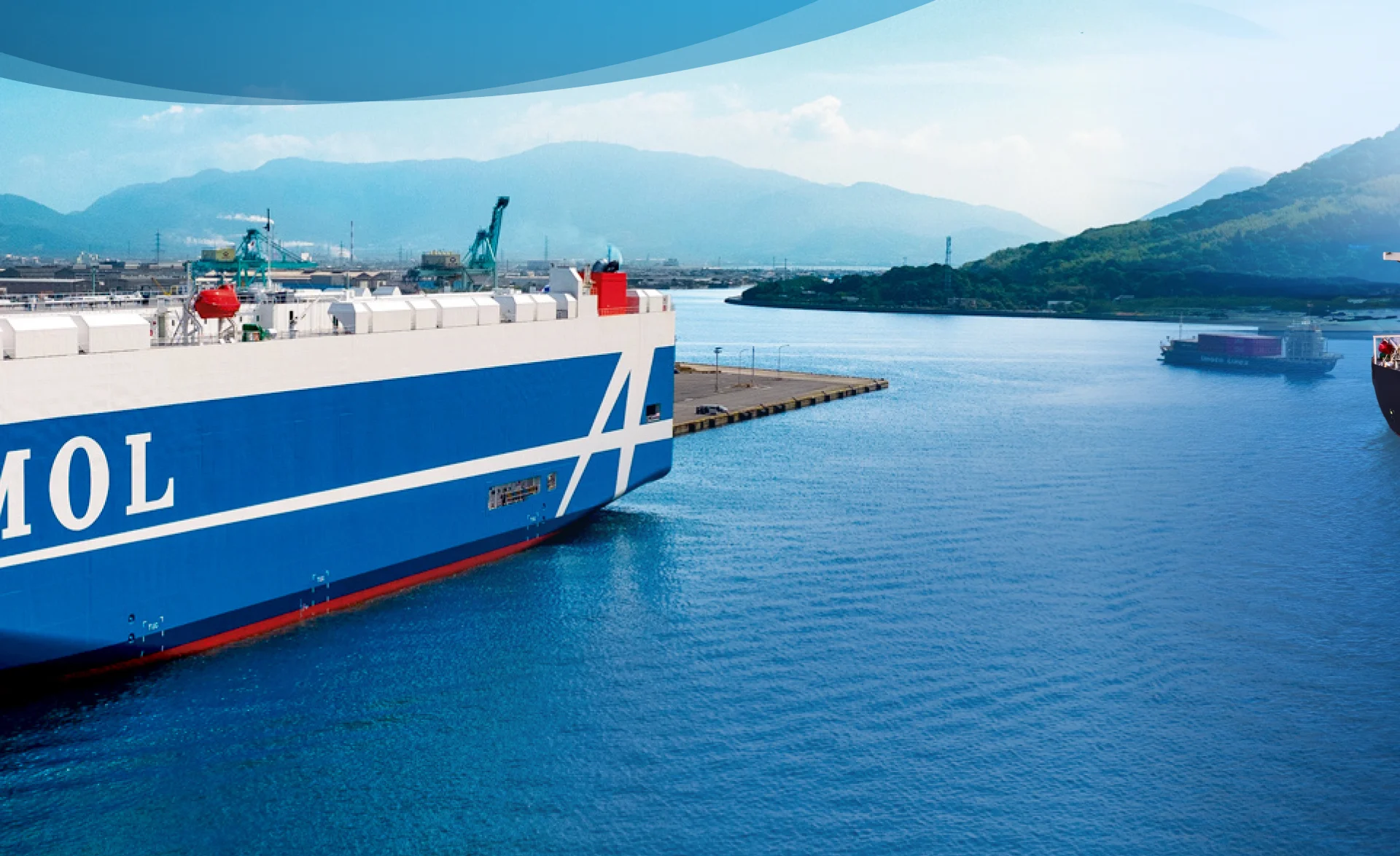 A large blue and white cargo ship is docked at a harbor near mountains under a clear sky, with calm blue water and another ship in the distance.