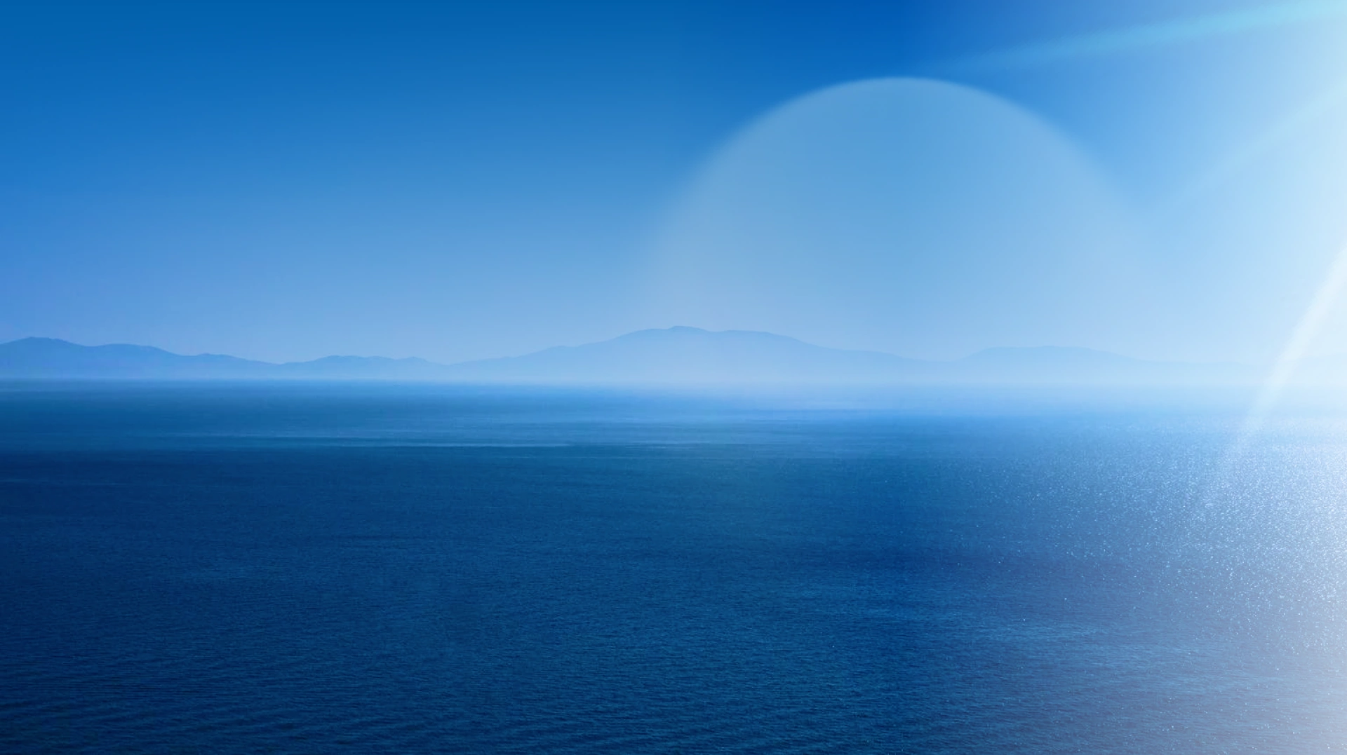A serene, deep blue ocean under a clear sky with faint mountainous outlines in the distance. Soft sunlight creates a peaceful, tranquil atmosphere.