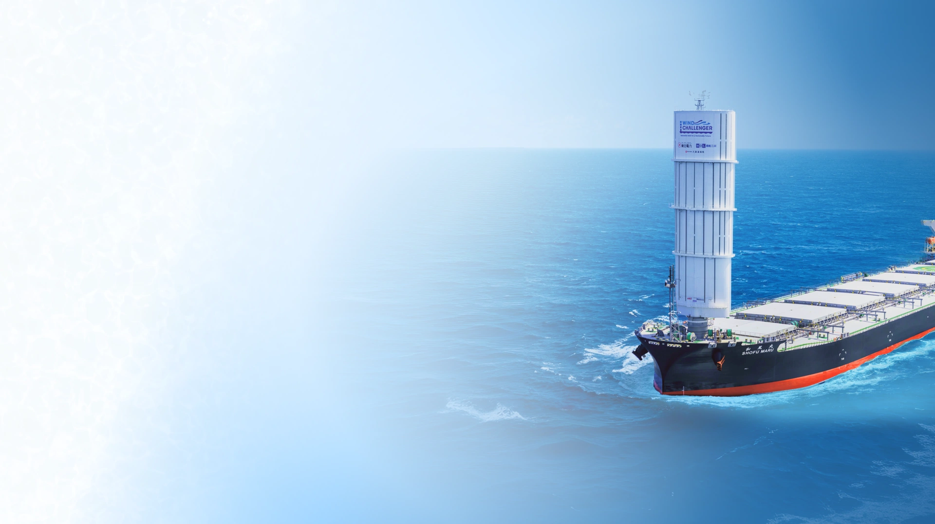 A cargo ship sails the ocean with a large white sail structure at the bow. The scene conveys innovation in maritime transport against a bright blue sea.