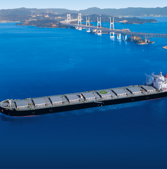 A large cargo ship sails through a vast, calm blue sea with a distant bridge and hilly landscape in the background, conveying tranquility and scale.