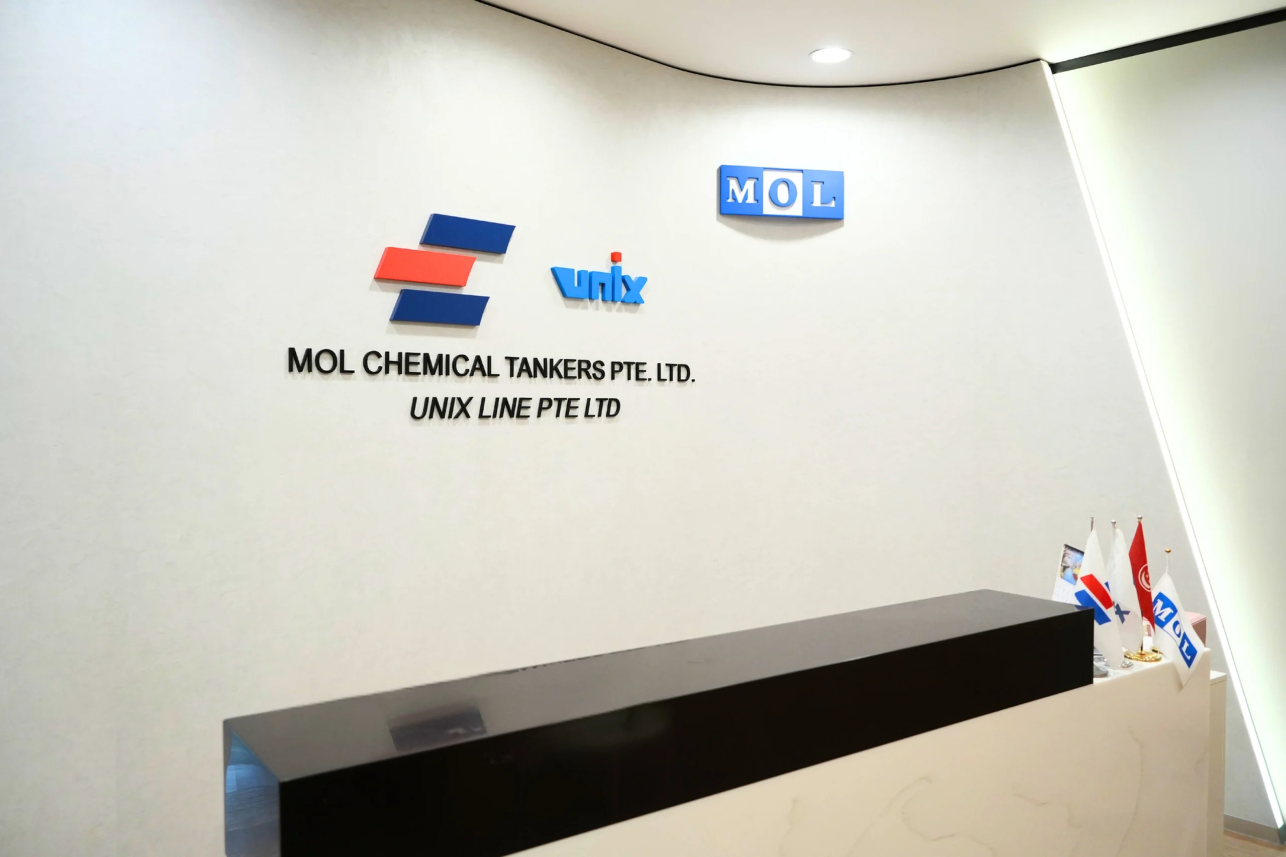 Reception area with "MOL Chemical Tankers Pte. Ltd." logo on a white wall. Minimalist decor with small flags and bright overhead lighting.