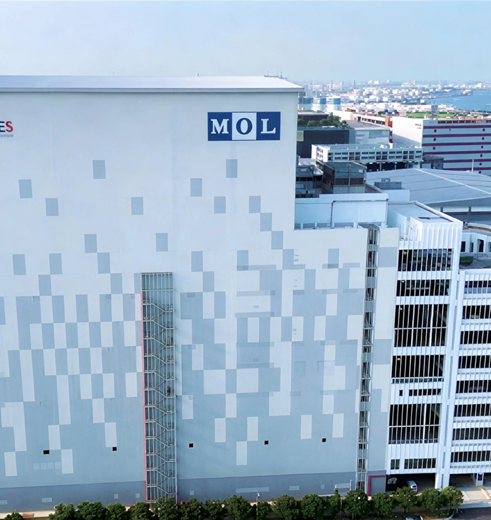A large industrial building with a white and gray pixelated facade displays the "MOL" logo. In the background, a harbor and urban landscape are visible under a clear blue sky.