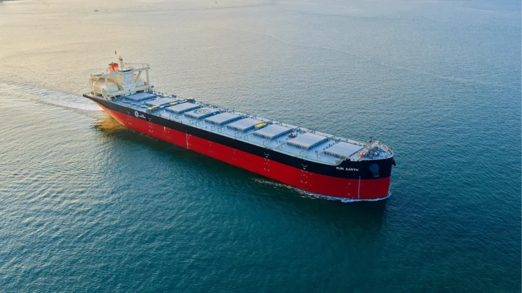 A large red and black cargo ship, part of the MOL Singapore Sub-Headquarters fleet, sails through calm blue water, viewed from above at an angle. The vessel features multiple covered holds and a white superstructure at the stern.