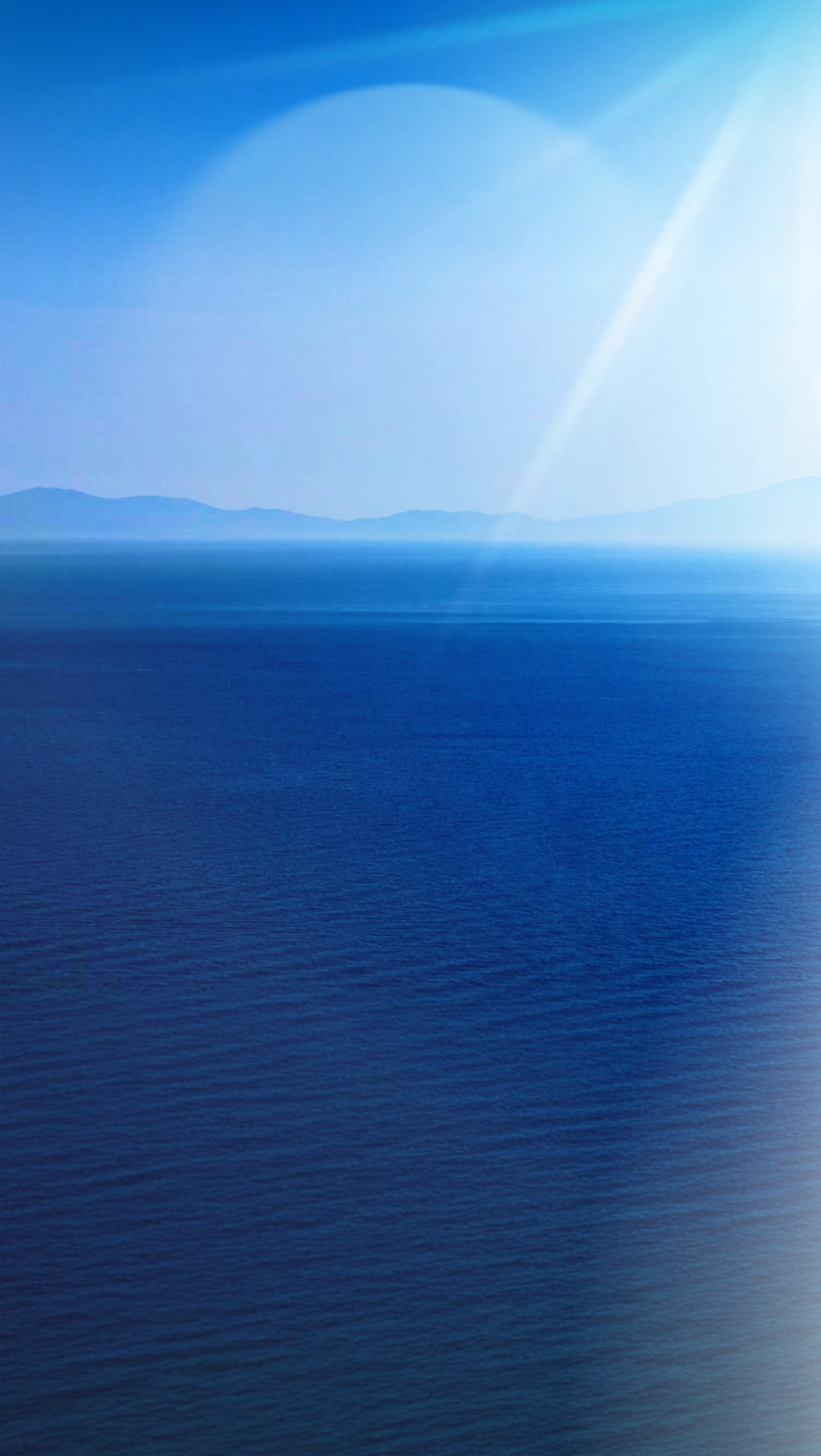 A calm blue sea stretches into the distance beneath a clear sky, with hazy mountains on the horizon and a beam of sunlight shining down from the upper right corner.
