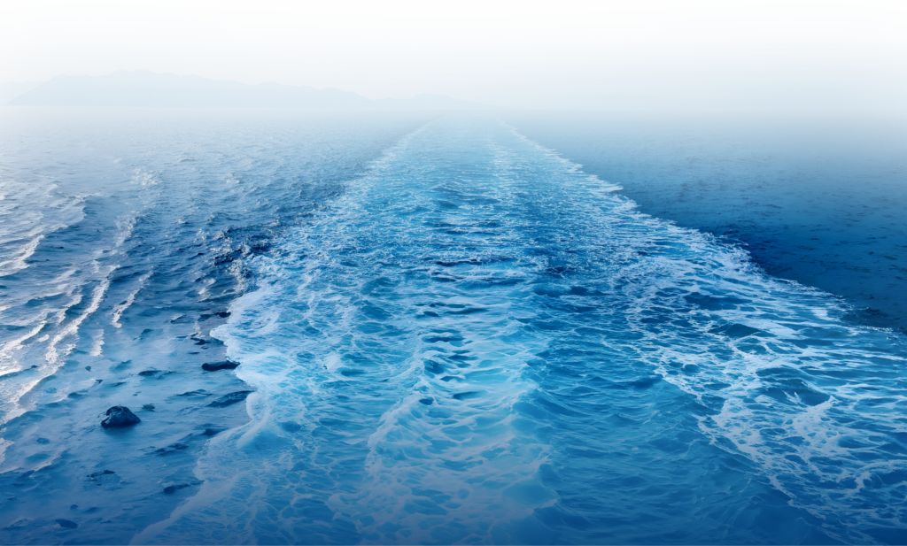 A wide trail of foamy blue water stretches back through a calm sea, fading into a misty, distant horizon where faint, hazy outlines of mountains rise—an inspiring vista reminiscent of MOL Group Southeast Asia & Oceania’s expansive reach.